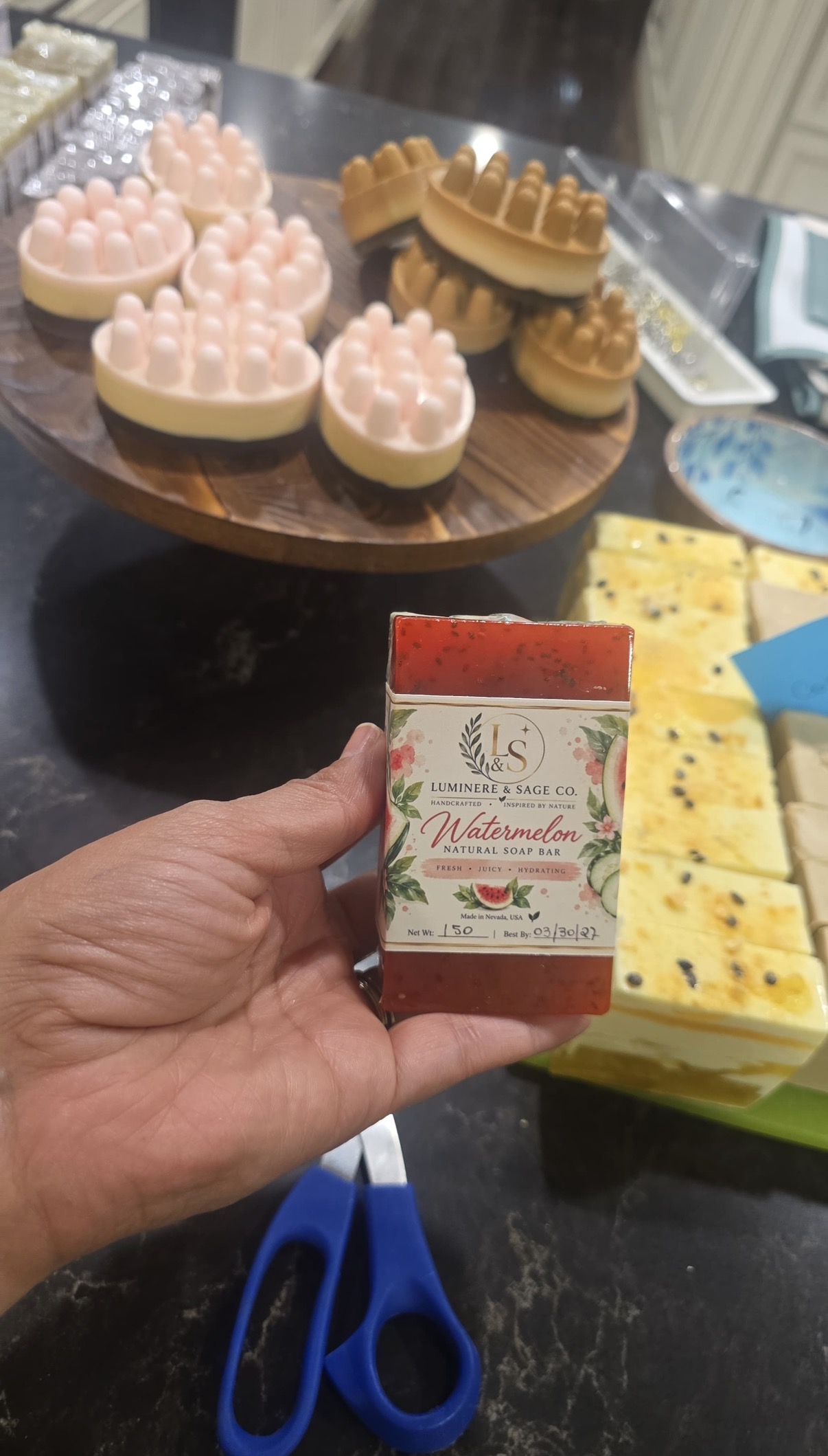Hand holding a wrapped red soap bar labeled Watermelon, with massage-shaped soaps behind