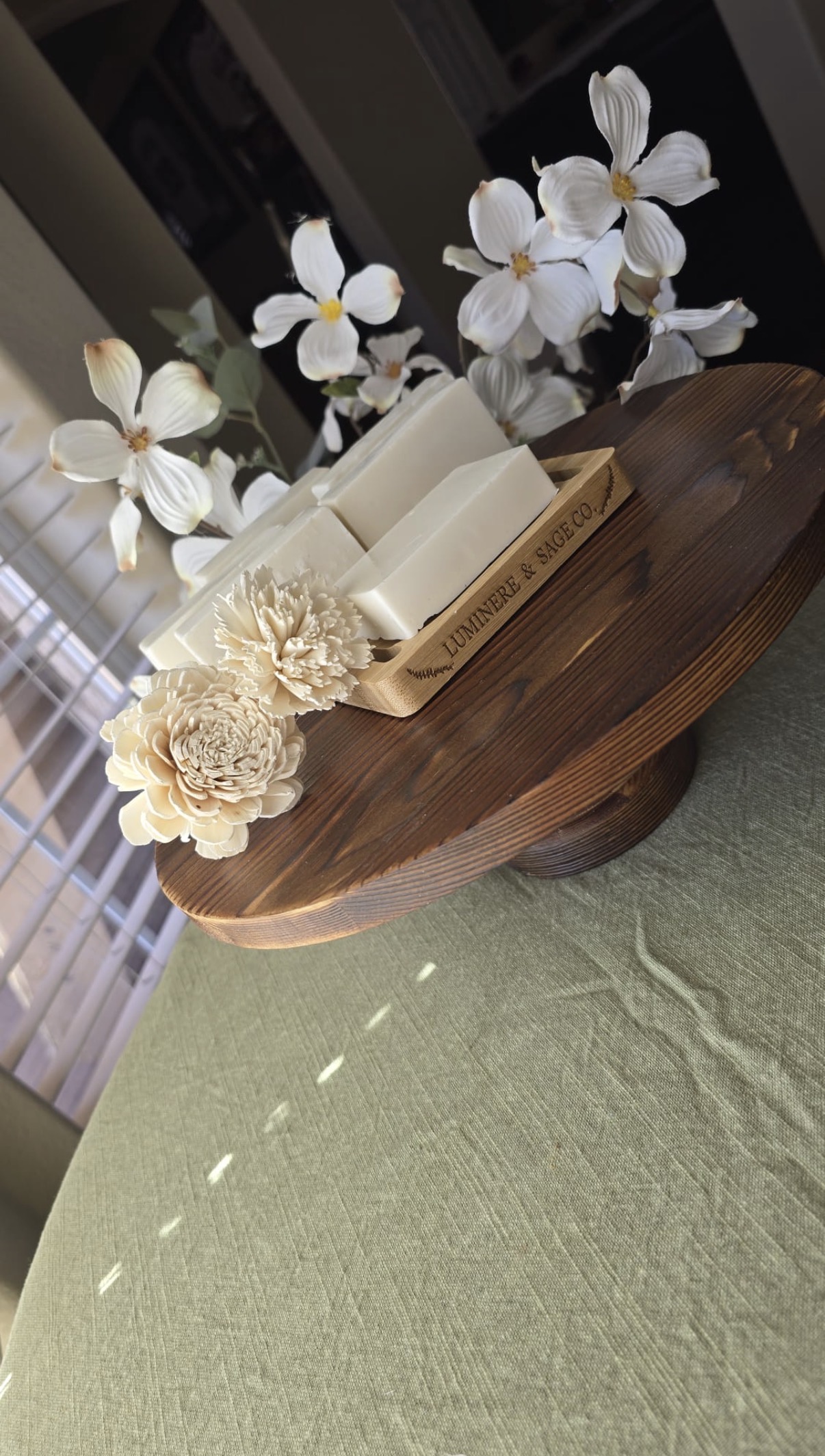 White rectangular soap bars on a branded bamboo tray with white flowers and cream floral shapes near a window