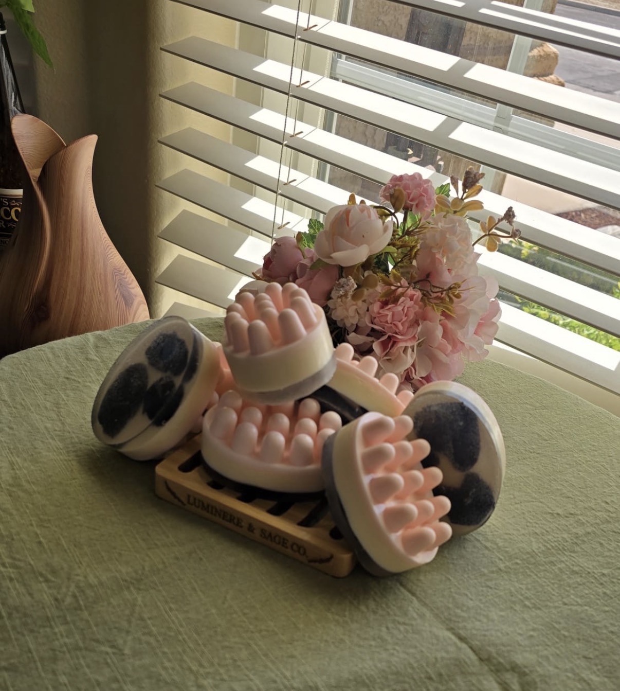 Pink, cream, and dark layered facial soaps on a branded tray with flowers near a window