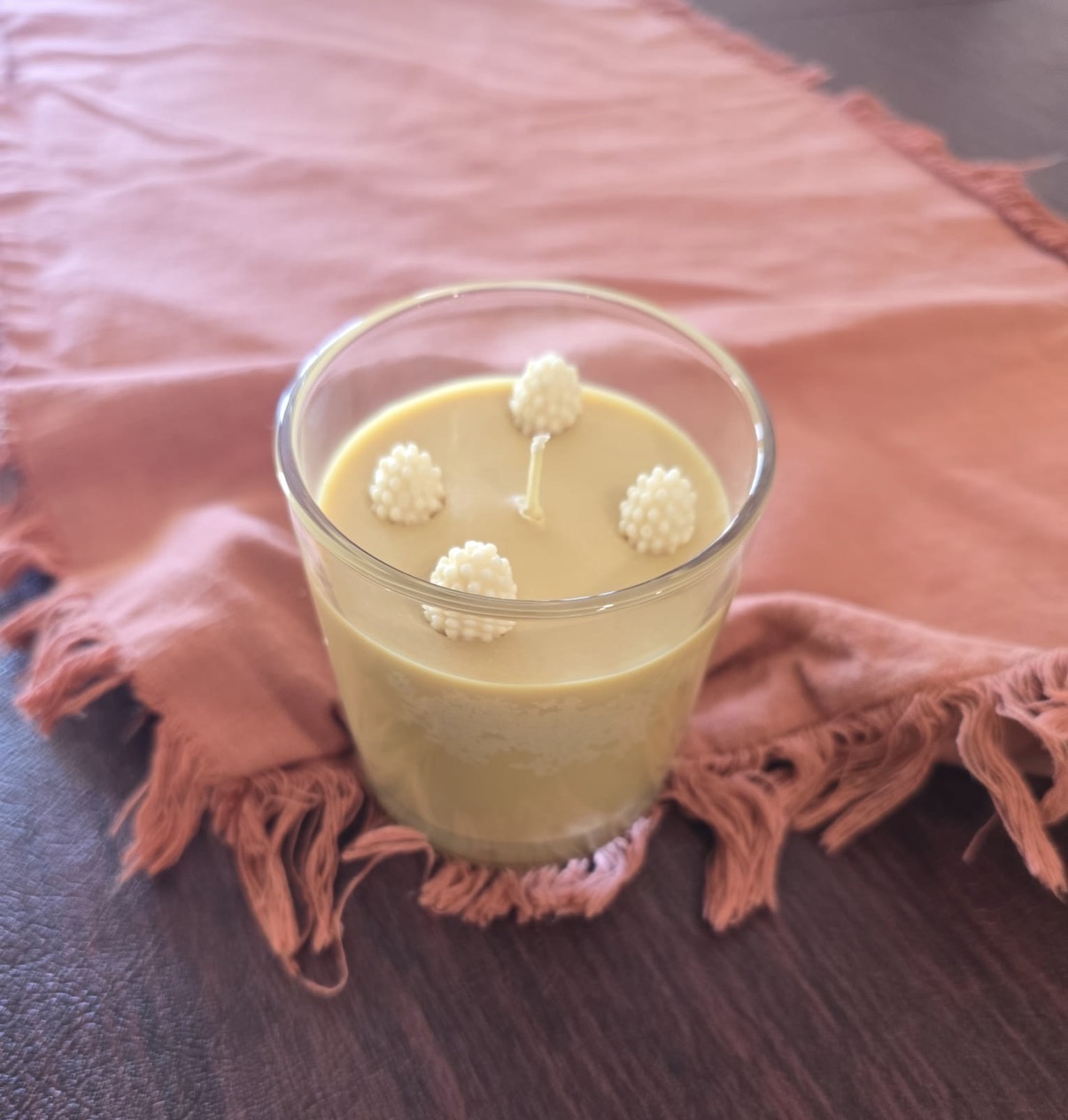 Pale cream and yellow candle in a clear glass with four white berry-shaped decorations and one unlit wick on pink cloth
