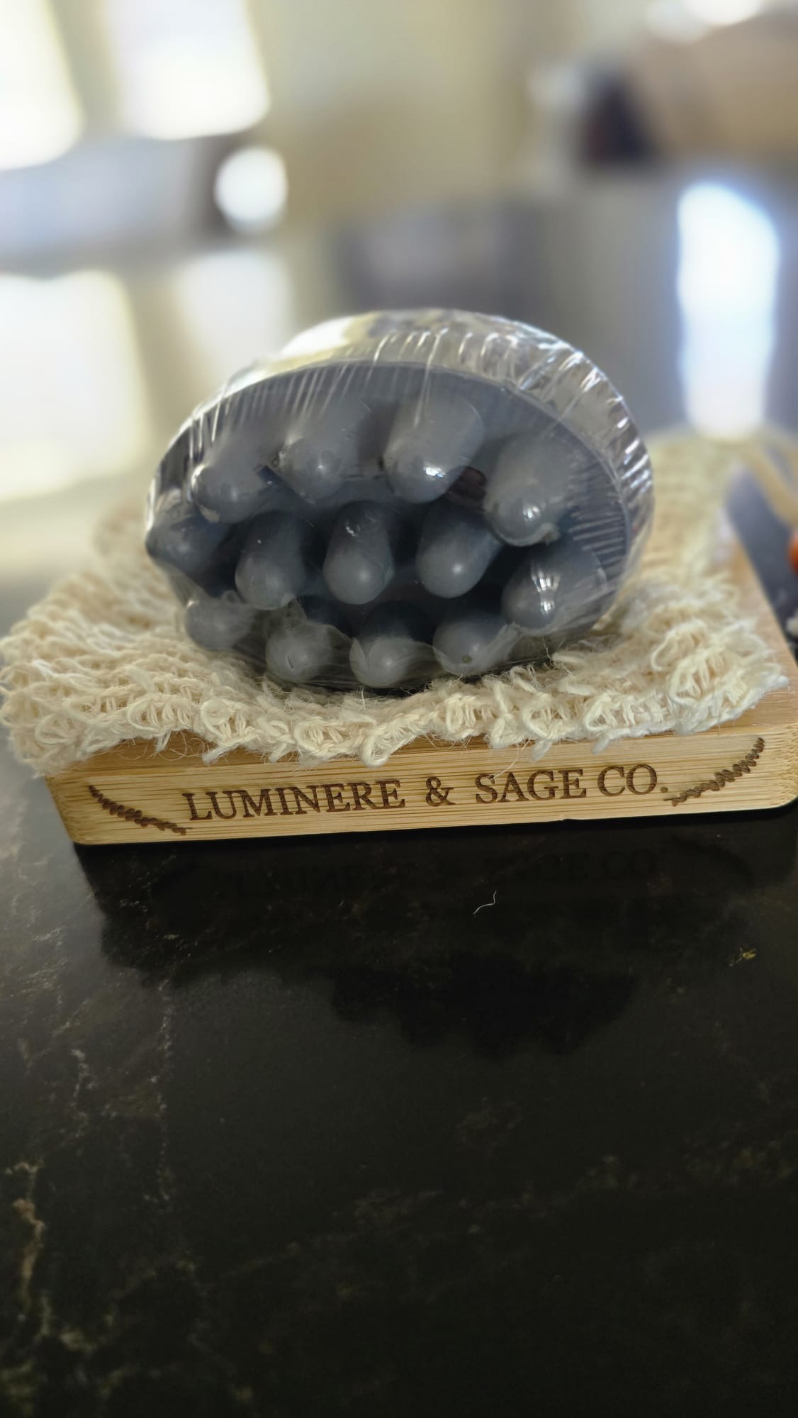 Wrapped dark gray massage-shaped facial soap on a cream pouch and Luminere & Sage branded bamboo tray